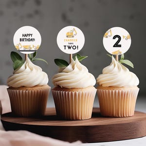 Puede incluir: Tres cupcakes con glaseado blanco y adornos decorativos. Los adornos dicen "Happy Birthday!", "Chadwick Turns Two!" y el número "2". Cada adorno presenta vehículos de construcción. Los cupcakes están sobre una tabla de madera.