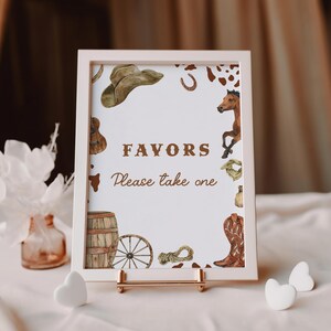 Op de afbeelding: Een ingelijst bord met een witte rand, met het woord "FAVORS" en de tekst "Please take one" in bruin. Het bord is versierd met aquarel illustraties van cowboy-thema-elementen, waaronder een hoed, hoefijzer, paard en laarzen.