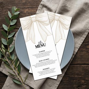 Könnte beinhalten: Zwei weiße Menükarten mit einer beigen Schleifenillustration werden auf einem grauen Teller präsentiert, mit dem Text "the MENU" oben. Die Speisekarte listet Vorspeisen, Hauptgerichte und Desserts auf. Ein Zweig grüner Blätter befindet sich links, und ein graues Tuch befindet sich rechts.