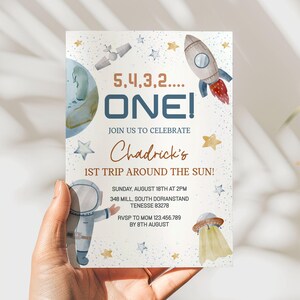 May include: A space-themed invitation with the text "5,4,3,2.... ONE!" and "Chadrick's 1st Trip Around the Sun!" features illustrations of a rocket, planet, stars, and an astronaut. The invitation is held in a hand.