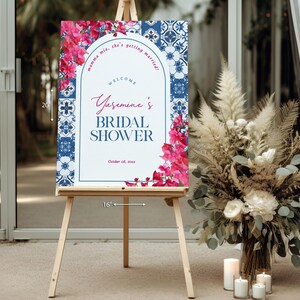 May include: A wooden easel displays a 20-inch by 16-inch bridal shower sign. The sign features a blue and white tile pattern border, pink floral accents, and the text "Yasemine's Bridal Shower".