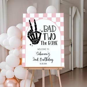 May include: A party sign on an easel with a pink and white checkered background. The sign reads "BAD TWO The BONE" with a skeleton hand making a peace sign. Below the text is "Welcome to Sabrina's 2nd Birthday 10.28.20XX". White and rose gold balloons are in the background.