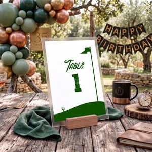 Może przedstawiać: Ustawienie stołu z numerem stołu w tematyce golfa. Znak jest zielono-biały, ze słowem "Table" i numerem "1". Czarny baner głosi "Happy Birthday". Inne elementy to kieliszek do koktajli, kubek i zegarek kieszonkowy.