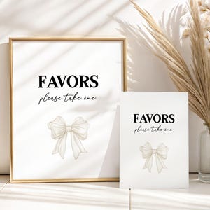 May include: Two white signs with the text "FAVORS please take one" in black script. The signs feature a watercolor bow illustration. One sign is framed in gold. The signs are for a wedding or event.