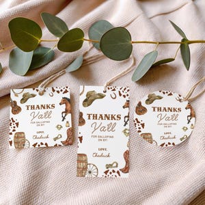 Op de afbeelding: Drie cadeaulabels met een western thema. Elk label heeft de tekst "Thanks Y'all" en cowboy-thema illustraties, waaronder een paard, hoed en laarzen. De labels zijn vastgebonden met touw.