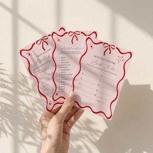 May include: Pink bridal shower game cards with red decorative borders and bows. The cards feature the text "How Old Was the Bride-to-Be?" and "Bridal Word Scramble". The cards are held in a hand.