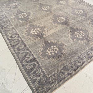 May include: A rectangular gray rug with a geometric pattern. The rug features a border with a repeating design and a central field with stylized floral motifs in shades of gray and off-white. The rug is made of a woven material.