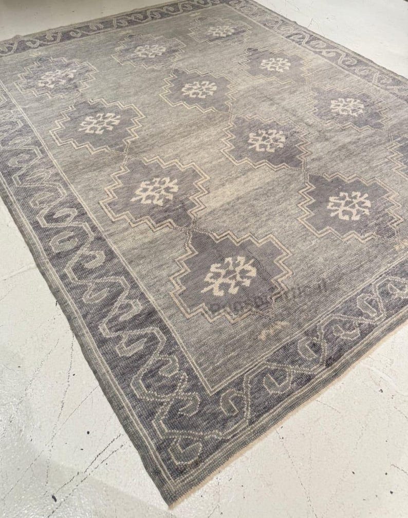 May include: A rectangular gray rug with a geometric pattern. The rug features a border with a repeating design and a central field with stylized floral motifs in shades of gray and off-white. The rug is made of a woven material.