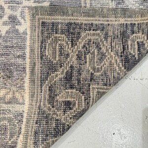 May include: Close-up of a section of a blue and gray patterned rug. The rug features a geometric design with a border of repeating patterns in darker shades of blue and gray. The rug is folded over, revealing the underside.