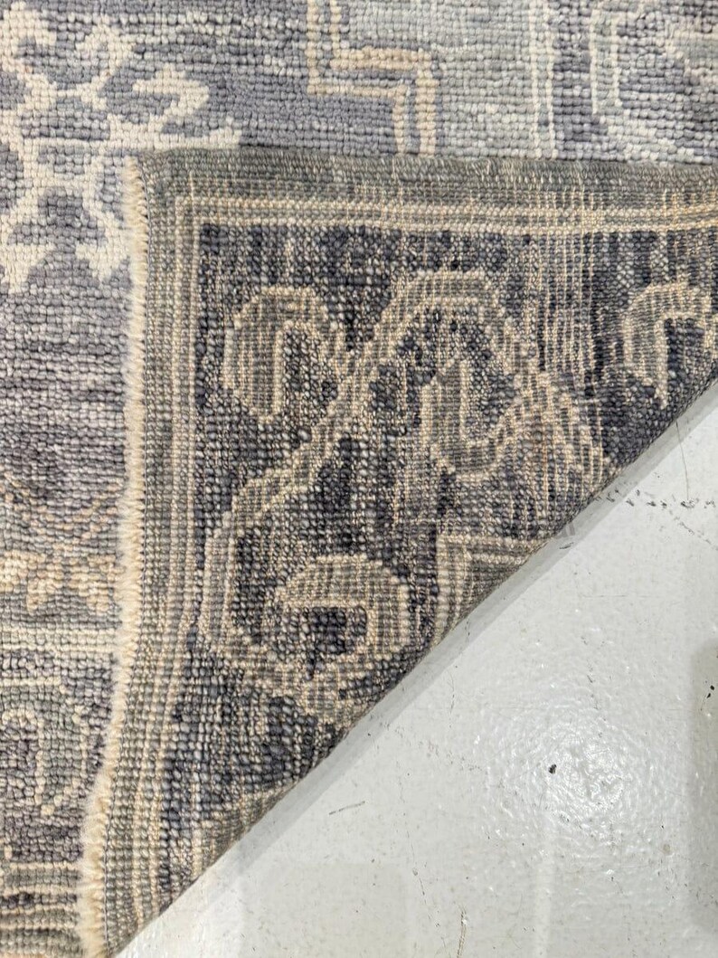 May include: Close-up of a section of a blue and gray patterned rug. The rug features a geometric design with a border of repeating patterns in darker shades of blue and gray. The rug is folded over, revealing the underside.