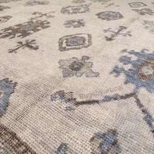 May include: Close-up of a beige rug with a repeating pattern of geometric shapes in shades of blue and brown. The rug has a textured surface and a border design. The text "Rugsbyartical" is visible on the rug.