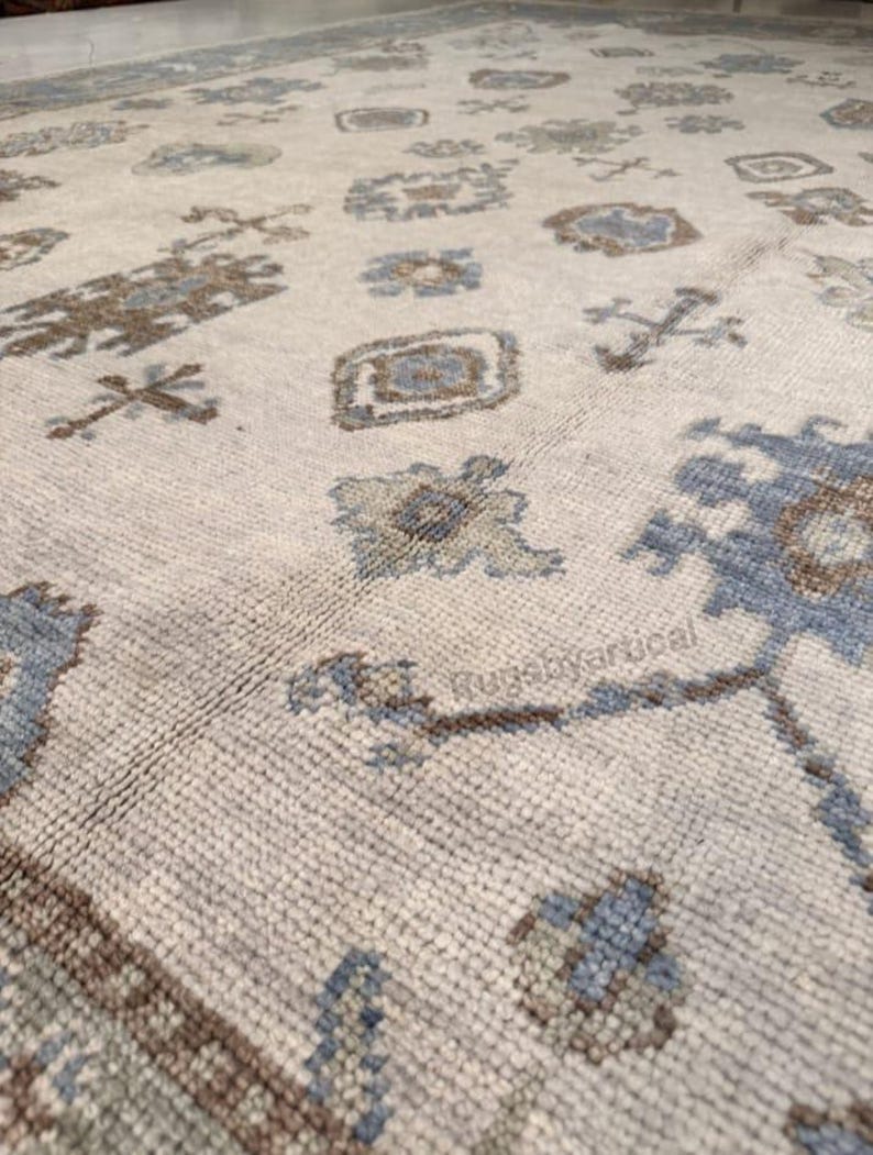 May include: Close-up of a beige rug with a repeating pattern of geometric shapes in shades of blue and brown. The rug has a textured surface and a border design. The text "Rugsbyartical" is visible on the rug.