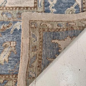 May include: Close-up of a section of a blue and beige patterned rug. The rug features a detailed floral design with a beige border. The corner of the rug is folded over, revealing the underside. The text "Rugsbyartica" is visible.