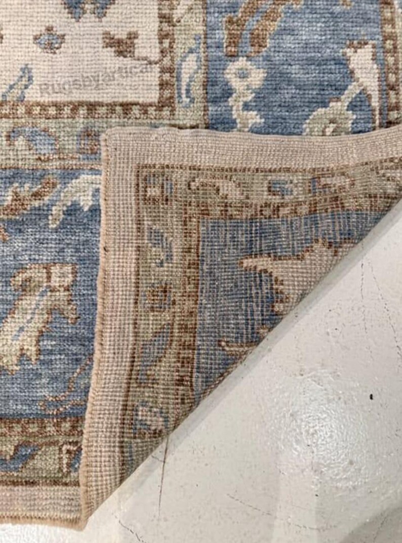 May include: Close-up of a section of a blue and beige patterned rug. The rug features a detailed floral design with a beige border. The corner of the rug is folded over, revealing the underside. The text "Rugsbyartica" is visible.