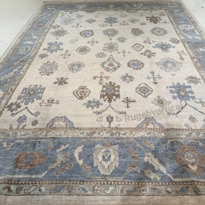 May include: A large rectangular area rug with a cream-colored field and a blue border. The rug features a repeating pattern of floral and geometric motifs in shades of blue, brown, and cream. The overall design is traditional and elegant.