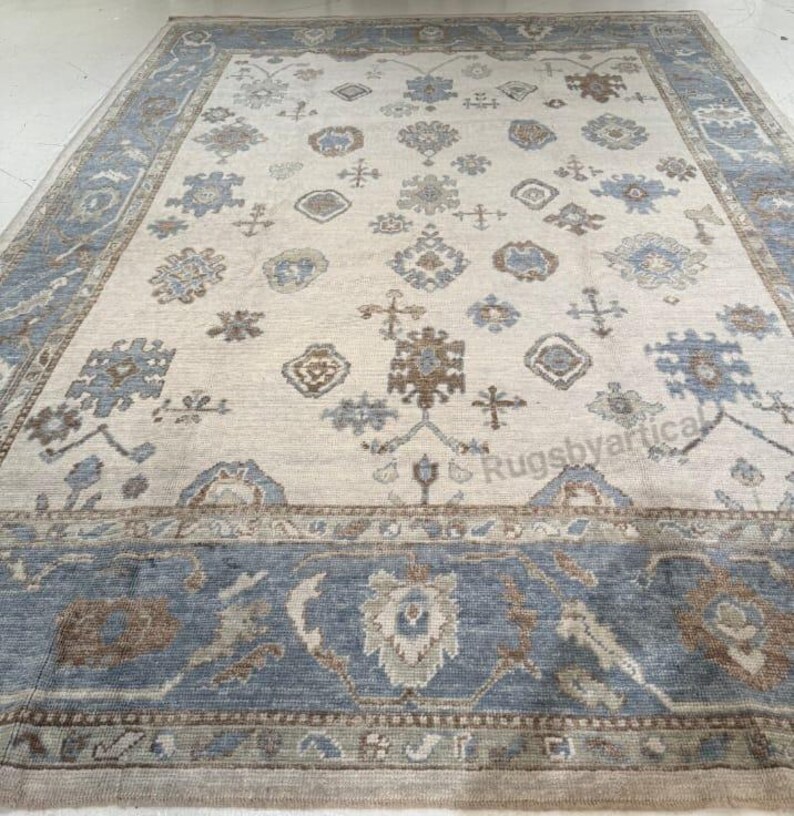 May include: A large rectangular area rug with a cream-colored field and a blue border. The rug features a repeating pattern of floral and geometric motifs in shades of blue, brown, and cream. The overall design is traditional and elegant.