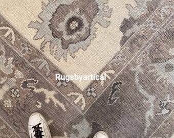 May include: A close-up shot of a beige and gray patterned rug with a floral design. The rug has a border with geometric shapes and the text "Rugsbyartical". White sneakers with black laces are visible at the bottom of the image.