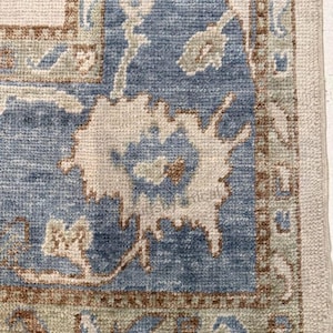 May include: Close-up of a section of a blue and beige rug with an intricate floral pattern. The rug features a central blue field with beige and brown accents, and a beige border. The design includes stylized floral motifs and geometric shapes.