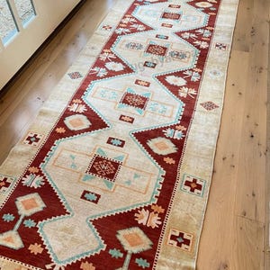May include: A long, rectangular rug with a central cream-colored field and a border of deep red. The rug features geometric patterns in shades of teal, orange, and cream. The rug is placed on a light-colored wooden floor.
