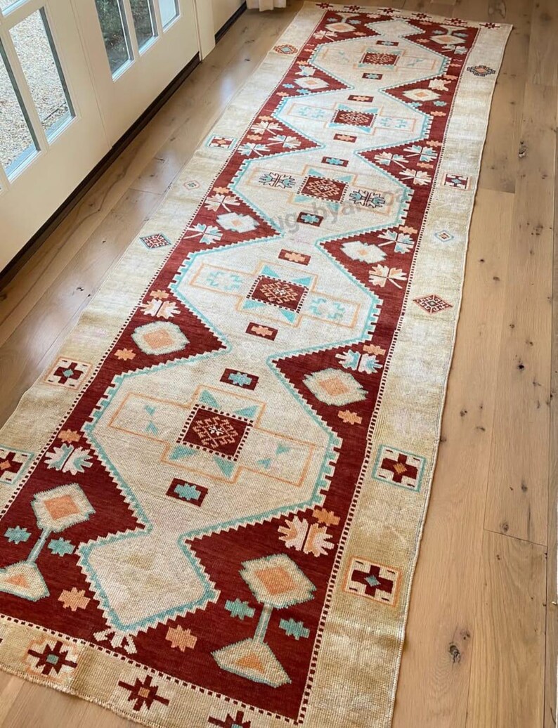 May include: A long, rectangular rug with a central cream-colored field and a border of deep red. The rug features geometric patterns in shades of teal, orange, and cream. The rug is placed on a light-colored wooden floor.