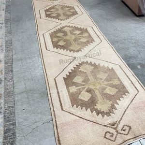 May include: A long, rectangular rug with a beige background and repeating geometric patterns in shades of brown and tan. The rug features a central design with a cross-like motif within a hexagonal frame, suitable for a hallway or entryway.