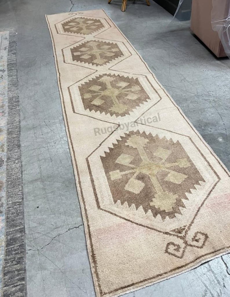 May include: A long, rectangular rug with a beige background and repeating geometric patterns in shades of brown and tan. The rug features a central design with a cross-like motif within a hexagonal frame, suitable for a hallway or entryway.