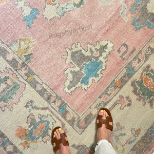 May include: A vintage-style rug with a pink central field and a patterned border. The rug features floral and geometric designs in shades of blue, orange, and gray. The text "Rugsbyvertical" is visible on the rug. Brown sandals and white pants are also visible.