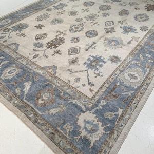 May include: A large, rectangular area rug with a cream-colored field and a blue border. The rug features a repeating floral pattern in shades of blue, brown, and gray. The border is a solid blue with a detailed design. The rug is made of a soft material.