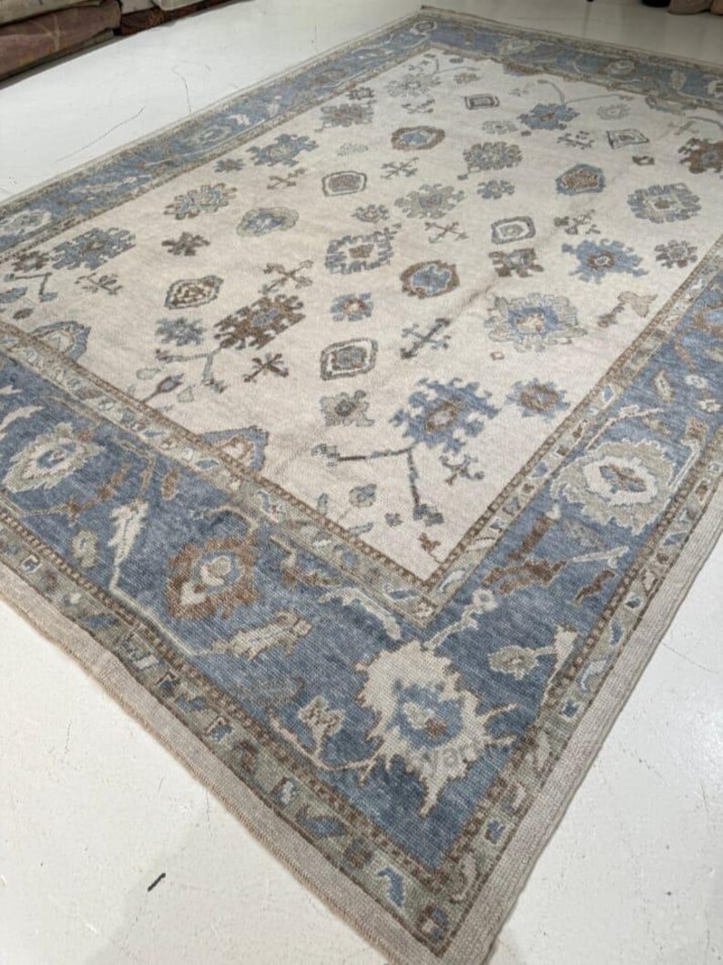 May include: A large, rectangular area rug with a cream-colored field and a blue border. The rug features a repeating floral pattern in shades of blue, brown, and gray. The border is a solid blue with a detailed design. The rug is made of a soft material.