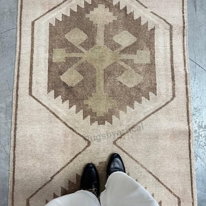 May include: A rectangular rug in beige tones, featuring a central geometric design in brown and olive green. The rug has a repeating pattern of diamond and zigzag shapes. The image includes a person's feet wearing black shoes and white trousers.