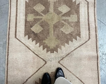 May include: A rectangular, beige-toned rug with a central geometric design in shades of brown and olive green. The rug features a repeating pattern of diamond and zigzag shapes. The image includes a person's feet wearing black shoes and white pants.