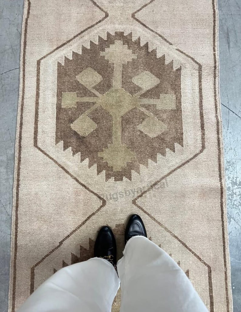 May include: A rectangular rug in beige tones, featuring a central geometric design in brown and olive green. The rug has a repeating pattern of diamond and zigzag shapes. The image includes a person's feet wearing black shoes and white trousers.