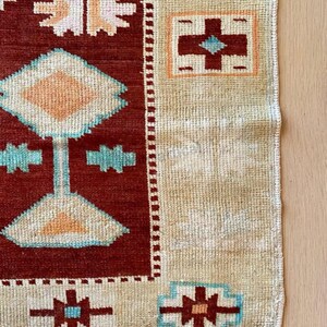 May include: A section of a handmade rug with a geometric pattern. The rug features a deep red central field with beige borders. The design includes diamond, cross, and star-like shapes in shades of beige, turquoise, and orange. The rug's texture is visible.