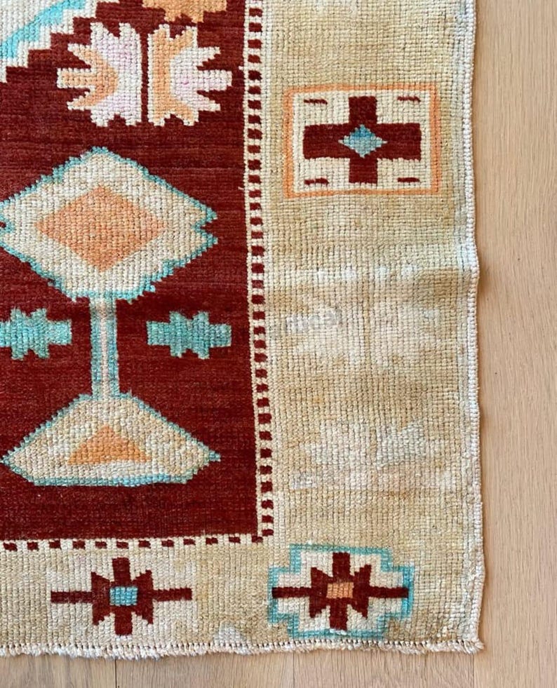 May include: A section of a handmade rug with a geometric pattern. The rug features a deep red central field with beige borders. The design includes diamond, cross, and star-like shapes in shades of beige, turquoise, and orange. The rug's texture is visible.