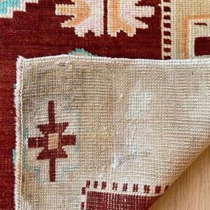 May include: Close-up of a section of a rug with a geometric pattern. The rug features a deep red border and central design, with accents of light blue, peach, and cream. The rug's underside is visible, showing a beige woven texture.