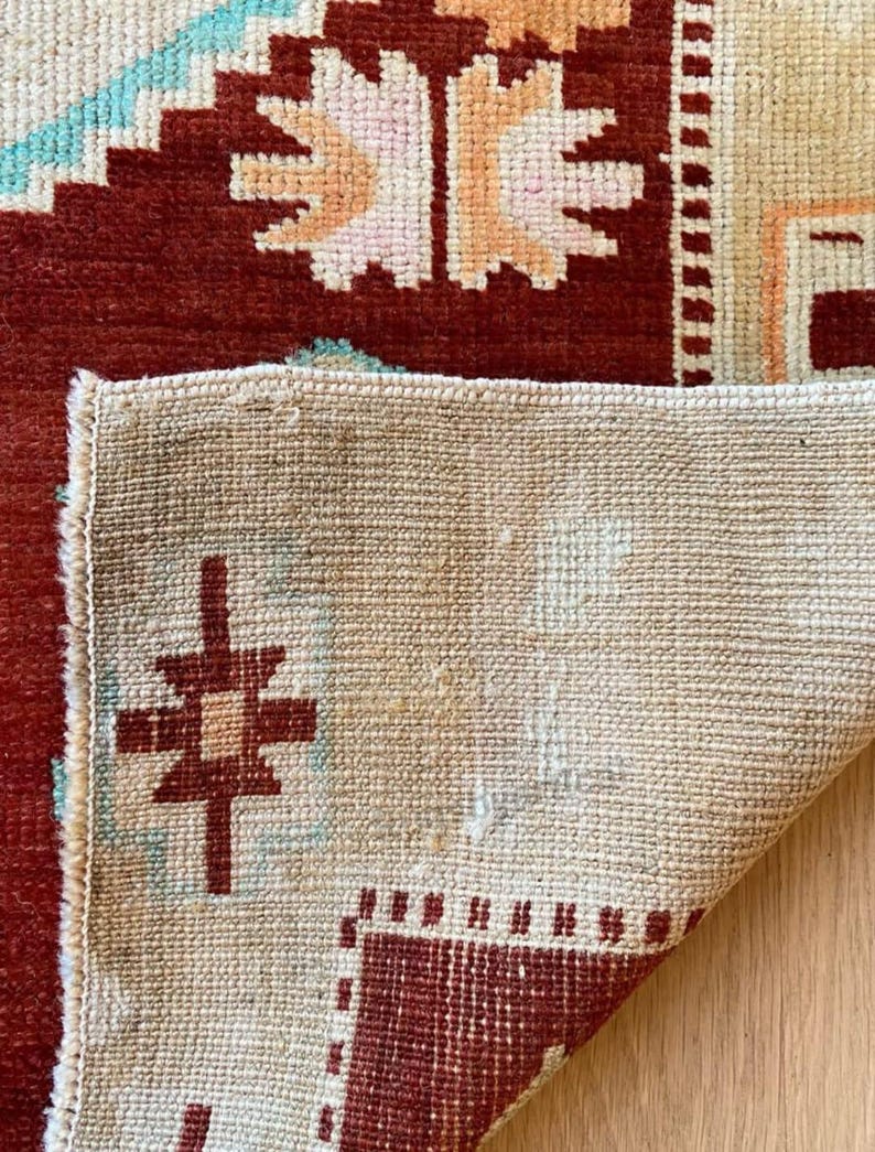 May include: Close-up of a section of a rug with a geometric pattern. The rug features a deep red border and central design, with accents of light blue, peach, and cream. The rug's underside is visible, showing a beige woven texture.