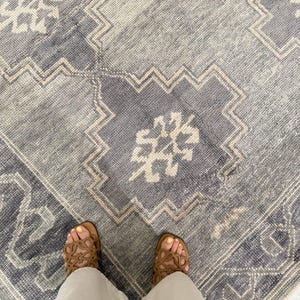 May include: A close-up view of a blue and beige patterned rug. The rug features a geometric design with a central medallion and a border. The rug's color palette includes shades of blue, beige, and white. The rug is made of a soft, textured material.