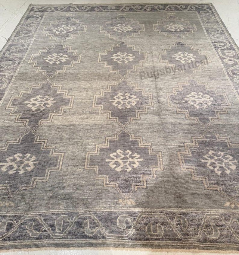 May include: A rectangular area rug in shades of gray and blue. The rug features a repeating geometric pattern of diamond shapes with floral accents. A border with a repeating design frames the rug.