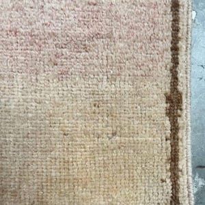 May include: Close-up of a beige rug with a rectangular shape. The rug has a textured surface with a faded pink pattern in the centre and a brown border. The rug is made of a soft material.