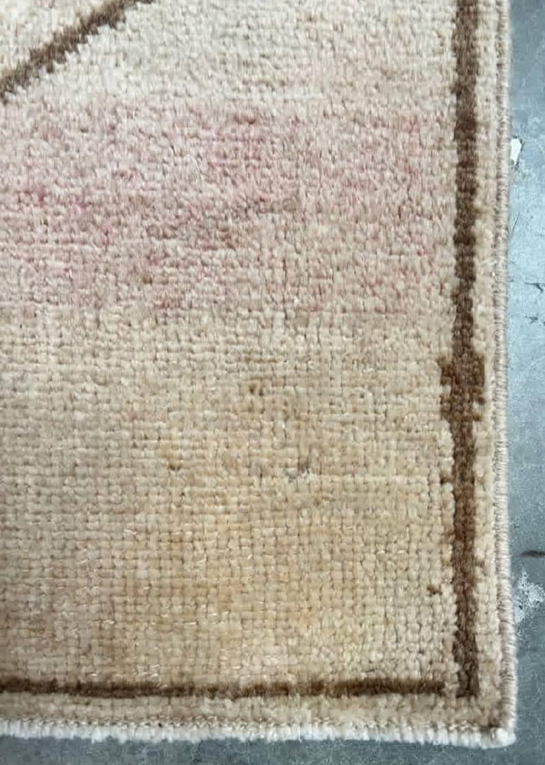 May include: Close-up of a beige rug with a rectangular shape. The rug has a textured surface with a faded pink pattern in the centre and a brown border. The rug is made of a soft material.