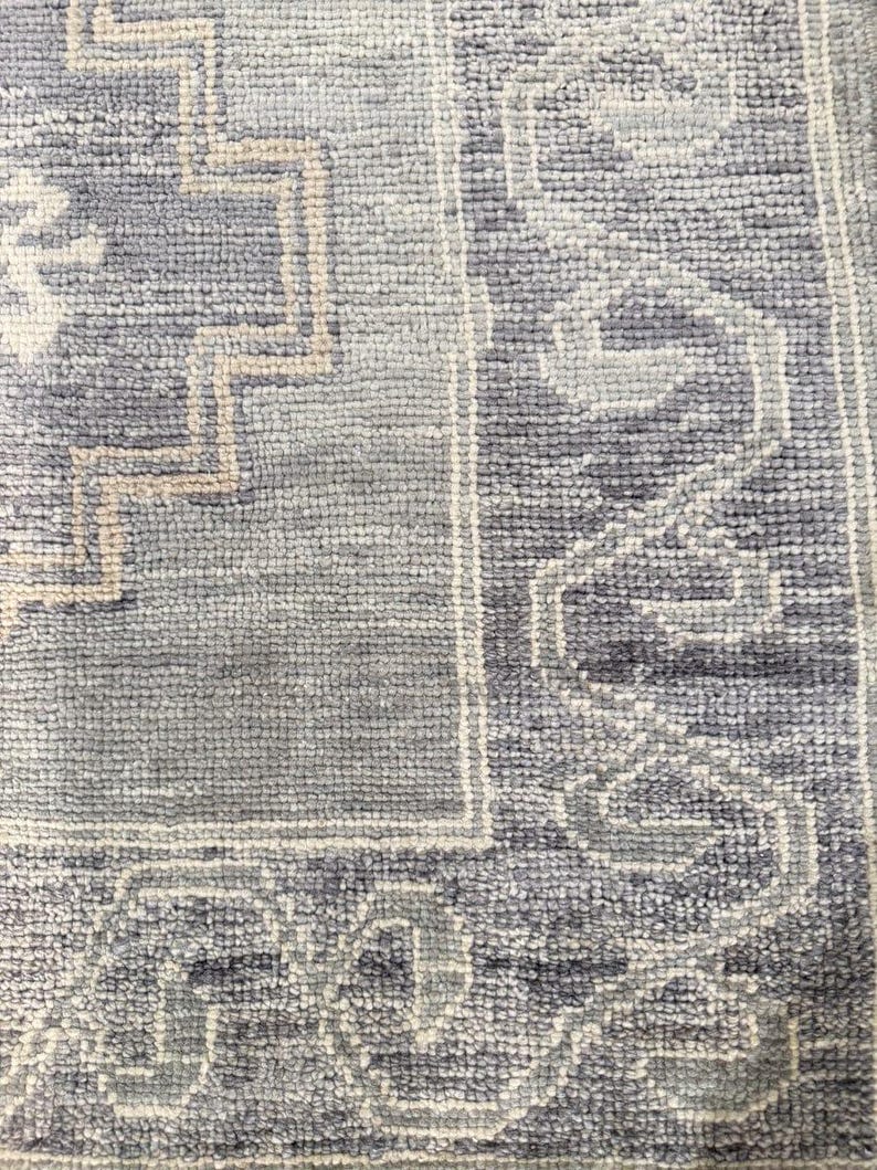 May include: Close-up of a textured, rectangular rug with a geometric pattern. The rug features a combination of light and dark blue hues, with beige and white accents. The design includes a border and central motifs.