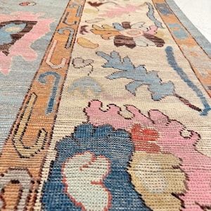 May include: A detailed shot of a vintage rug with a floral pattern. The rug features a light blue, beige, and orange color scheme with pink and blue floral designs. The rug has a long, rectangular shape.