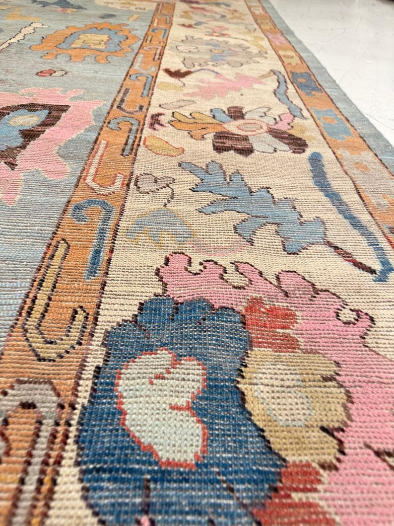 May include: A detailed shot of a vintage rug with a floral pattern. The rug features a light blue, beige, and orange color scheme with pink and blue floral designs. The rug has a long, rectangular shape.