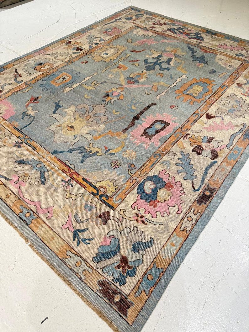 May include: A large, rectangular rug with a light blue central field and a beige border. The rug features floral and geometric patterns in shades of pink, orange, and brown. The rug is made of a woven material.