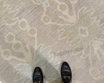 May include: A neutral-toned area rug with a floral pattern in shades of blue and green. The rug is partially folded, revealing the text "Rugsbycritical". Black leather loafers with gold accents are visible.