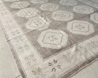 May include: A rectangular, neutral-toned rug with a repeating geometric pattern. The rug features a central field of muted brown with large, octagonal shapes in a lighter beige. A border with floral motifs frames the design.