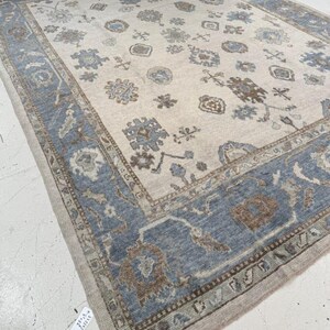 May include: A large, rectangular area rug with a cream-colored central field. The rug features a detailed border in shades of blue, brown, and beige. The central field is adorned with floral and geometric patterns in complementary colors, creating a traditional aesthetic.