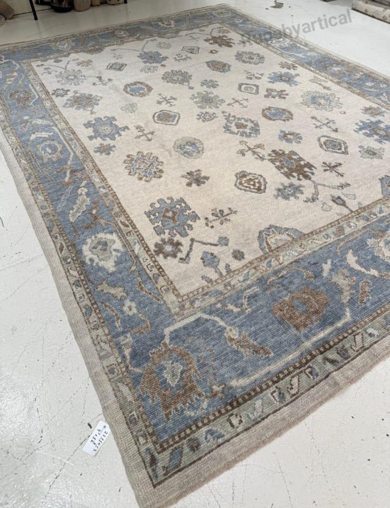 May include: A large, rectangular area rug with a cream-colored central field. The rug features a detailed border in shades of blue, brown, and beige. The central field is adorned with floral and geometric patterns in complementary colors, creating a traditional aesthetic.
