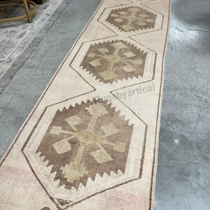 May include: A long, rectangular rug with a repeating geometric pattern. The rug has a cream-coloured background with brown and tan geometric shapes. The design is symmetrical, with the shapes aligned down the centre. The rug is likely made of woven material.