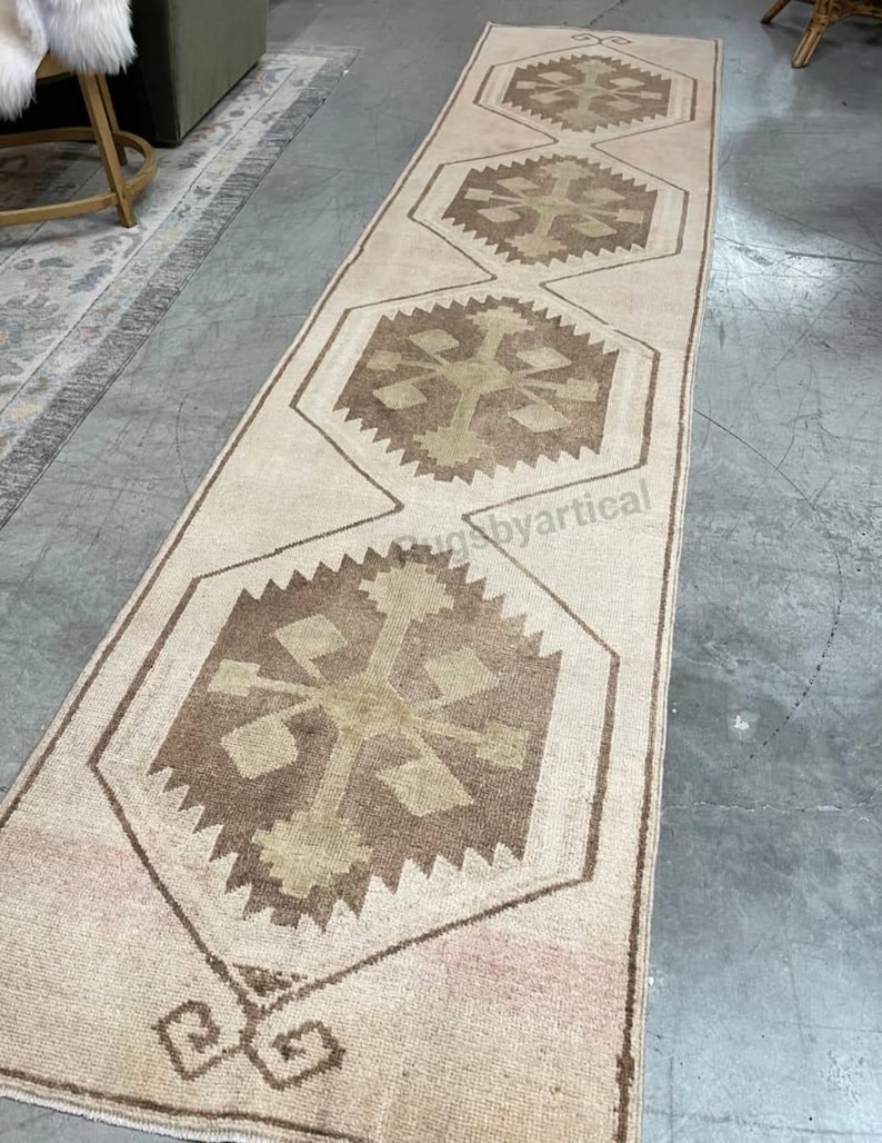May include: A long, rectangular rug with a repeating geometric pattern. The rug has a cream-coloured background with brown and tan geometric shapes. The design is symmetrical, with the shapes aligned down the centre. The rug is likely made of woven material.